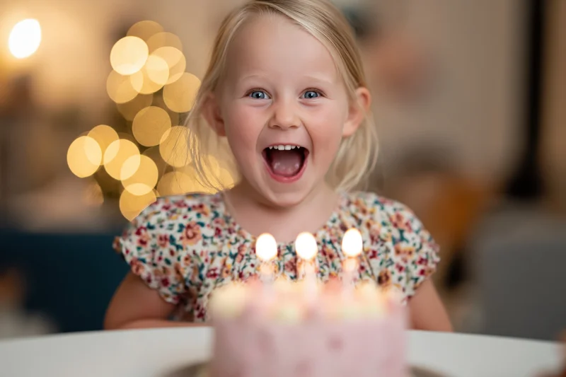 Excited young blonde girl with mouth open in front of birthday cake with lit candles at home