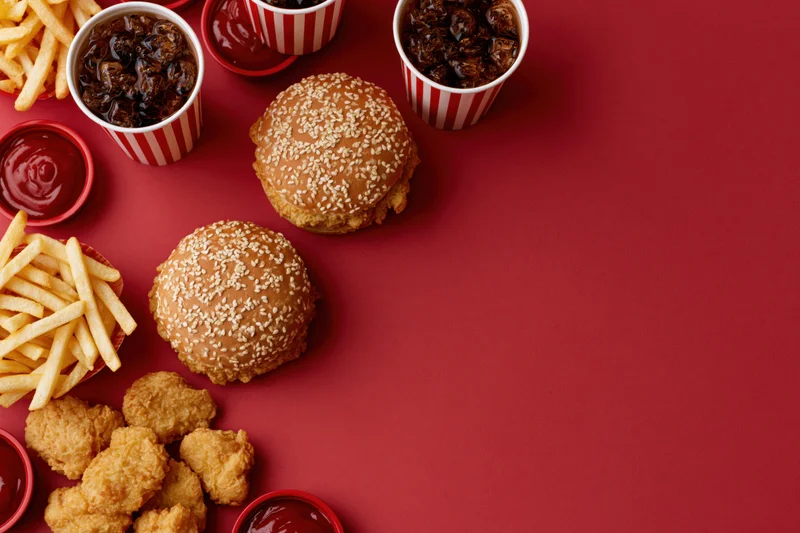 Fast food items including burgers, fries, chicken nuggets and ketchup arranged on a red surface in a flat lay composition