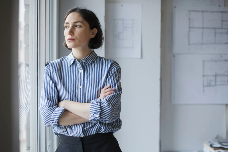 Female architect standing by studio window with arms crossed, gazing outside, blueprints on wall behind her