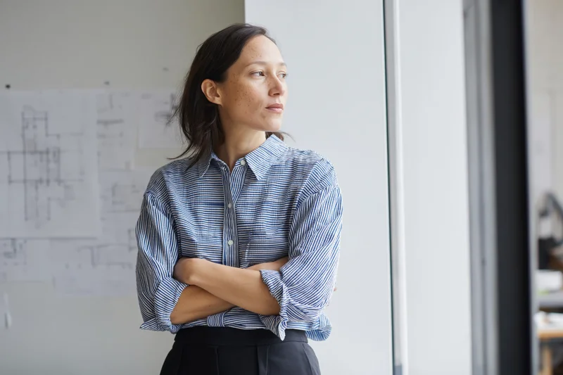 Female architect standing by a tall studio window with arms crossed, looking out, blueprints on the wall behind her