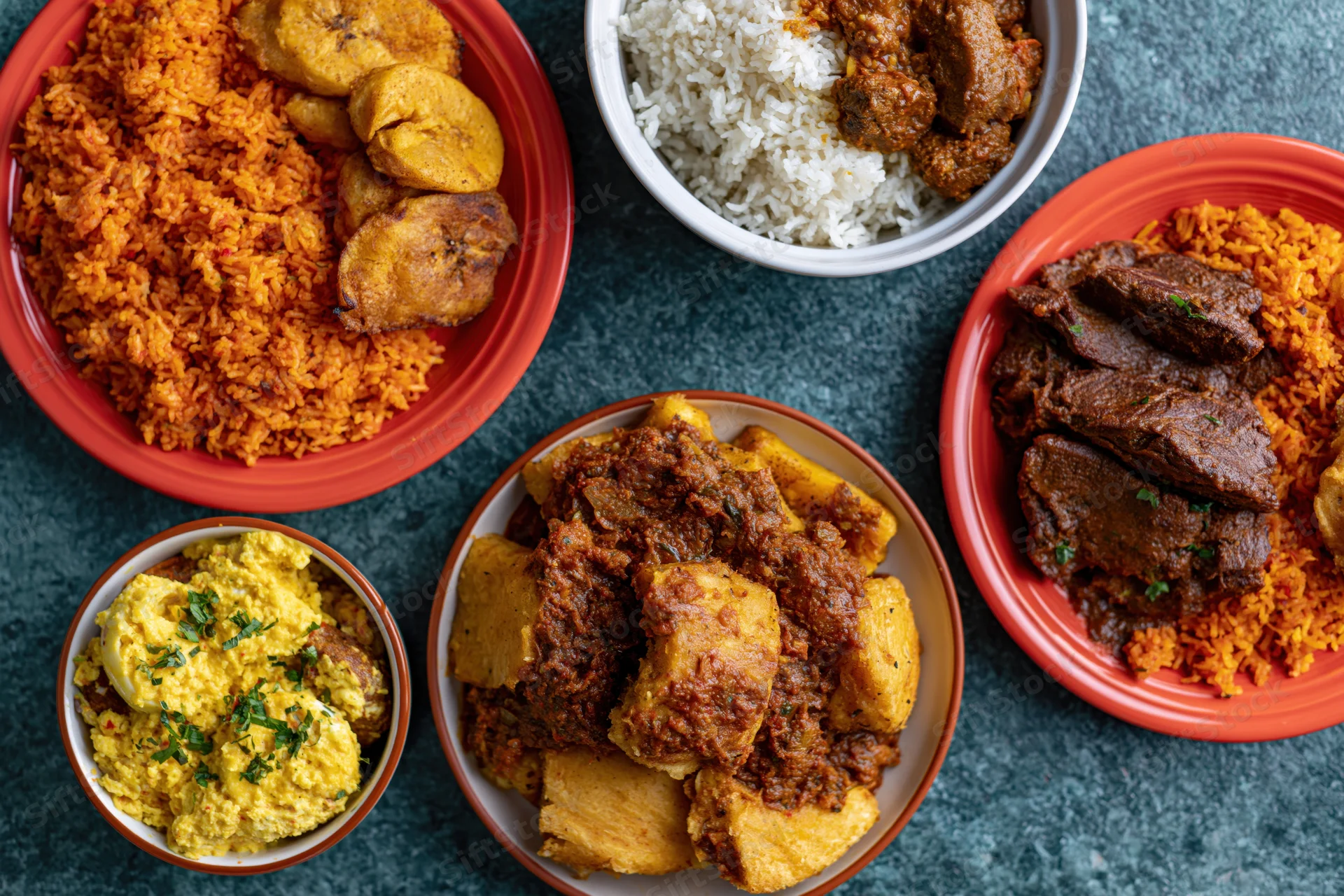 Five plates of west african food arranged on a dark teal surface with rice, stew, plantain and yam
