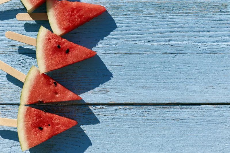 Watermelon slice popsicles on wooden sticks arranged on a blue painted wood surface in sunlight