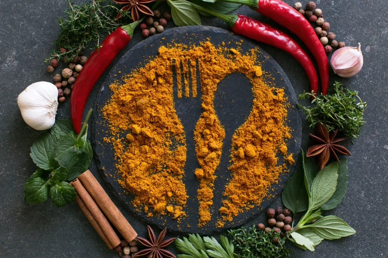 Turmeric powder shaped into cutlery on dark slate surrounded by herbs and spices