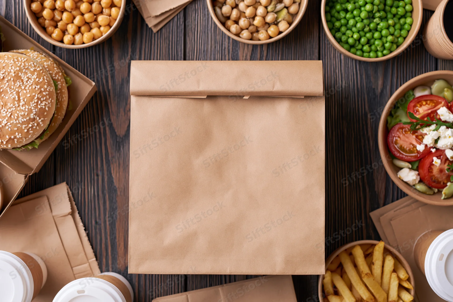 Kraft paper takeout bag centered on dark wood table surrounded by various food items and containers