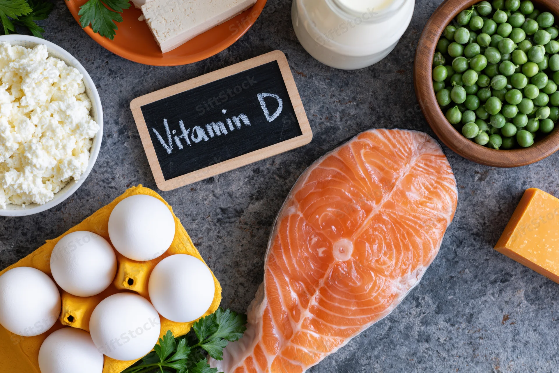 Salmon fillet, eggs, dairy products, tofu, peas, and cottage cheese arranged on dark grey surface with chalkboard sign reading vitamin d