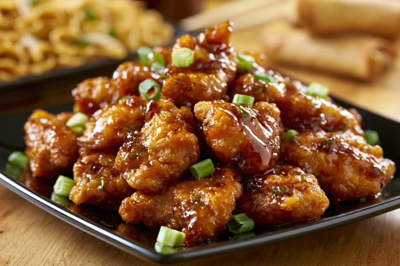 Glazed orange chicken pieces piled on a black square plate with green onions on a wooden table