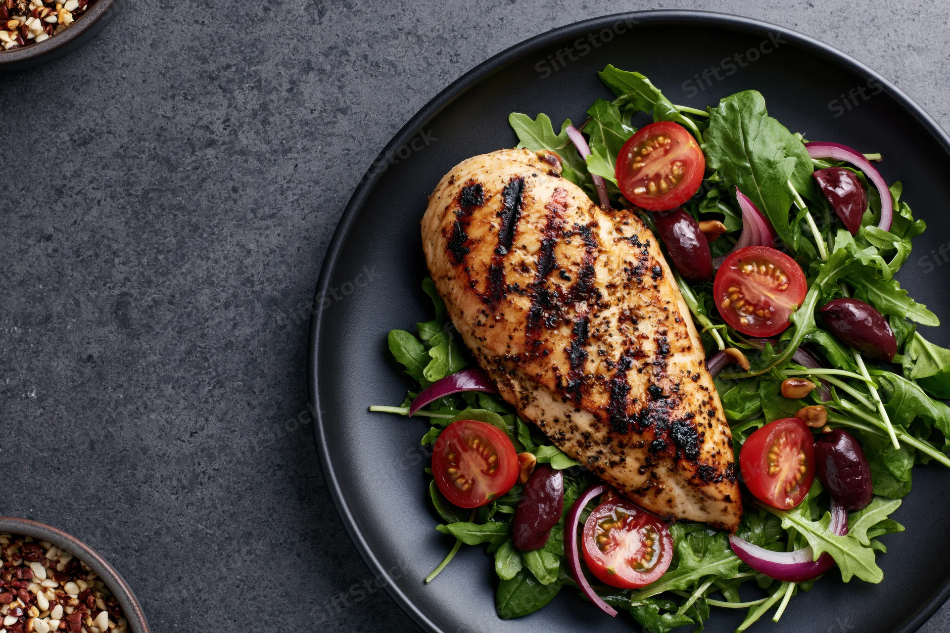 Grilled chicken breast with char marks on a bed of arugula salad with tomatoes, olives, and onions on a black plate