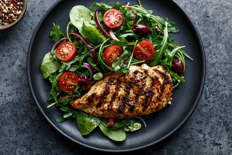 Grilled chicken breast with char marks served on a bed of arugula salad with tomatoes and olives on a black plate