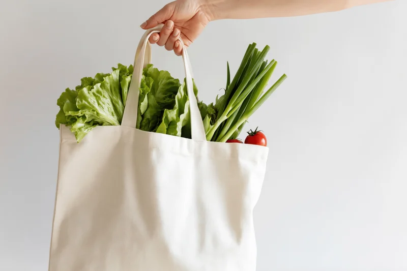 Hand holding a white canvas tote bag filled with fresh vegetables against a plain background