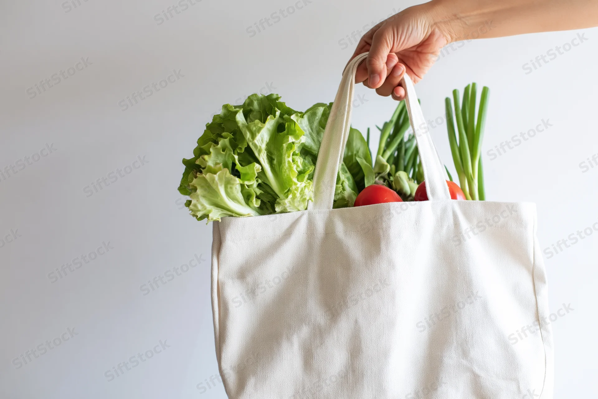 Hand holding a white canvas tote bag filled with fresh vegetables against a plain background