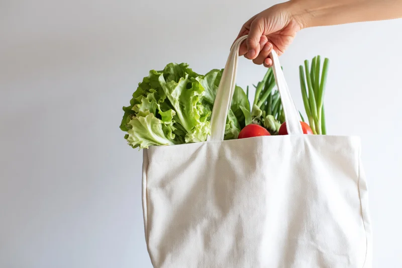 Hand holding a white canvas tote bag filled with fresh vegetables against a plain background