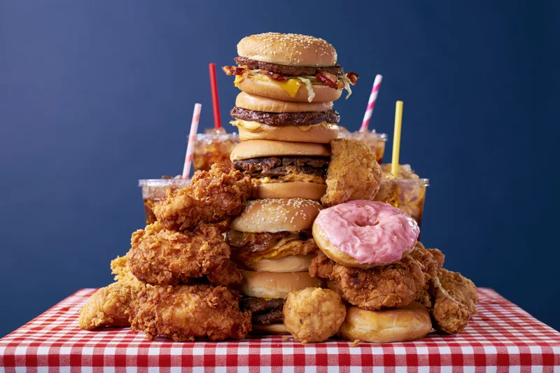 Towering stack of burgers, fried chicken, donuts and sodas on a red and white checkered tablecloth