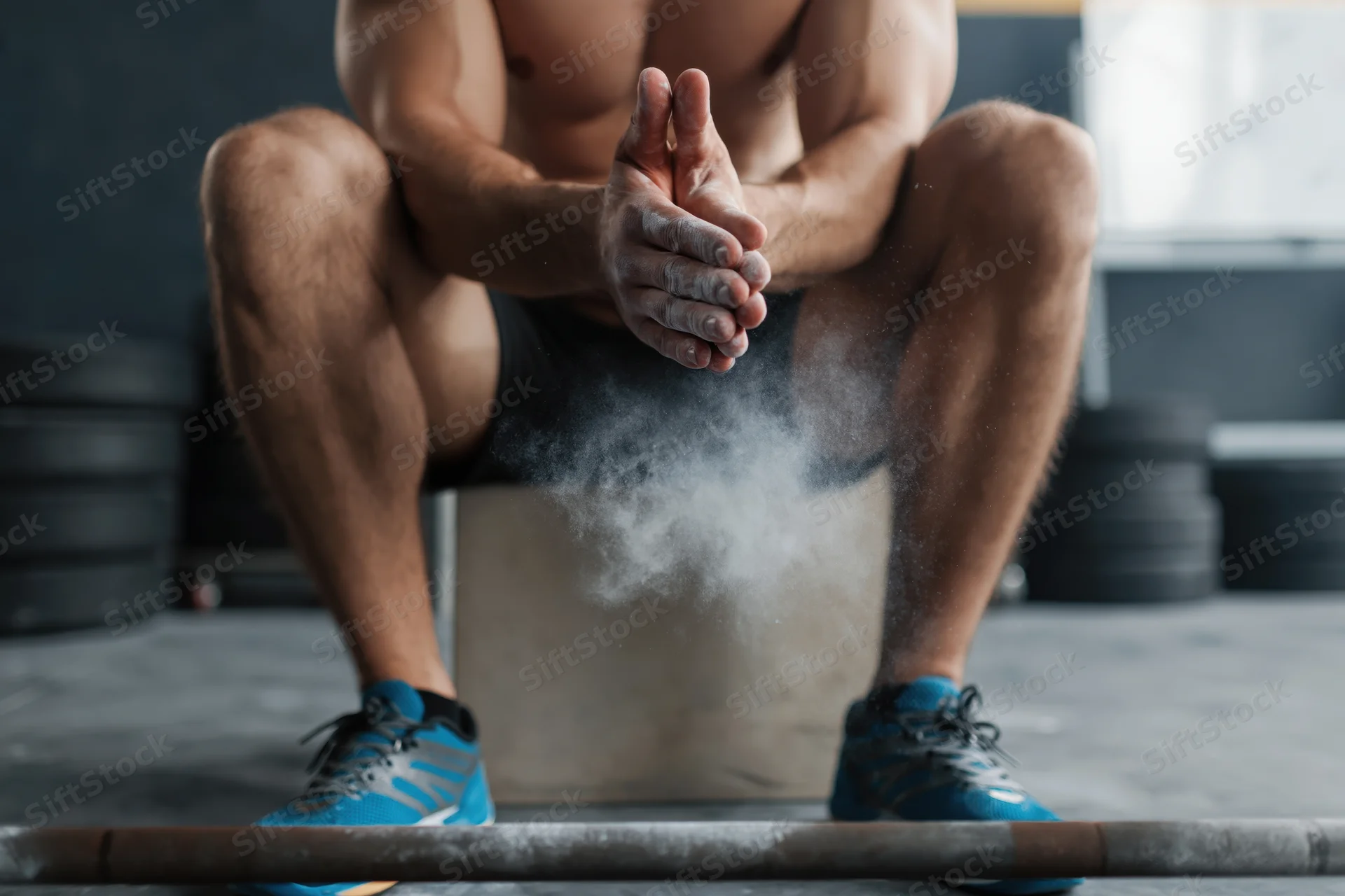 Male weightlifter sitting on box clapping hands with chalk dust rising near barbell in gym