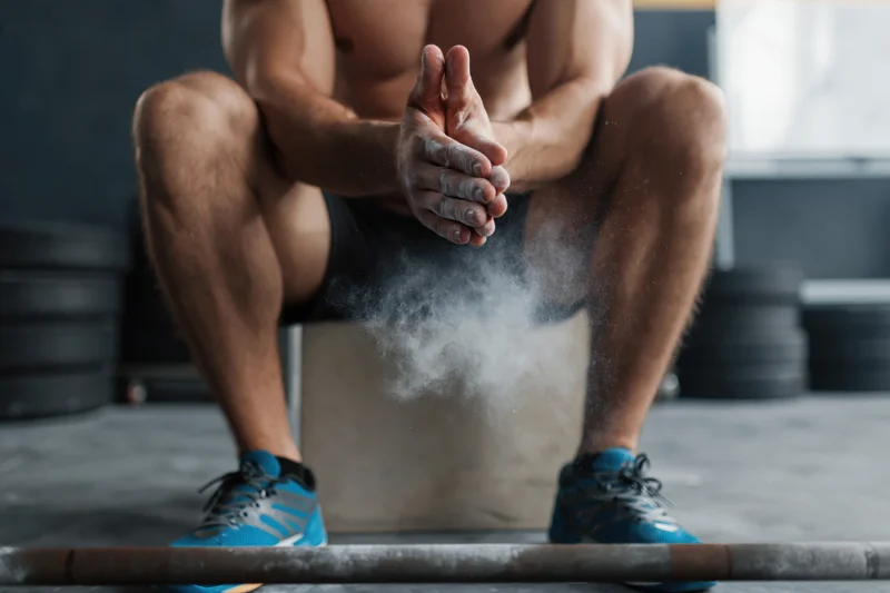 Male weightlifter sitting on box clapping hands with chalk dust rising near barbell in gym