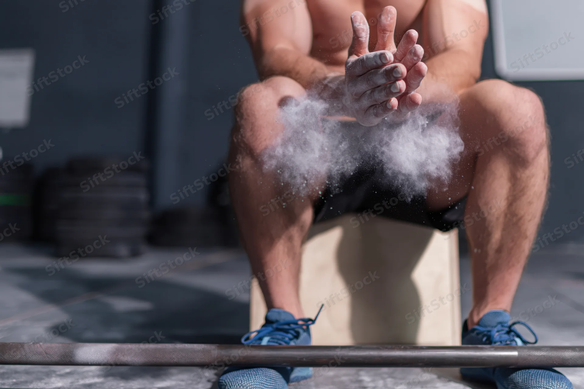 Male weightlifter sitting on wooden box clapping chalk-covered hands in gym with barbell in foreground
