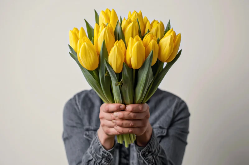 Man holding a large bouquet of yellow tulips in front of his face, wearing a dark grey denim shirt against a neutral background