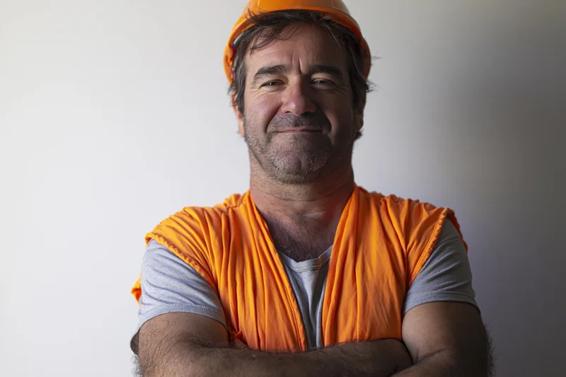 Man in orange safety vest and hardhat smiling with arms crossed against white background