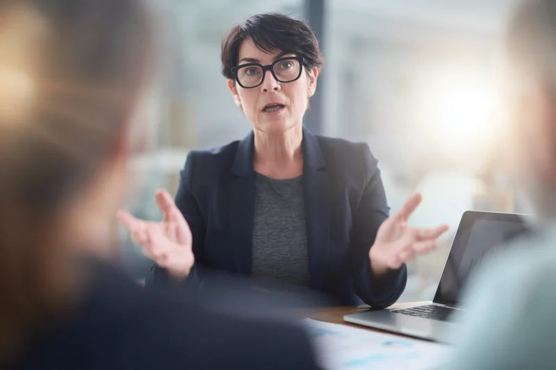 Mature businesswoman gesturing during meeting at office desk with laptop and documents