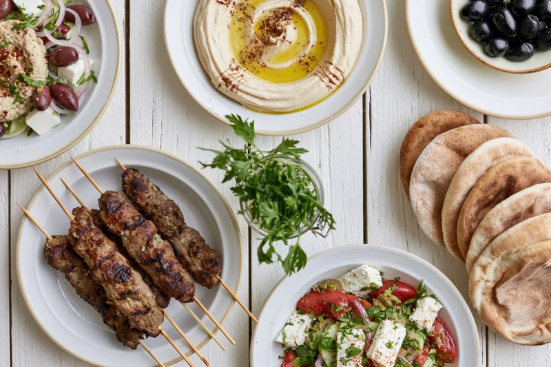 Mediterranean mezze spread with hummus, kebabs, pita, salad and olives on white wooden table