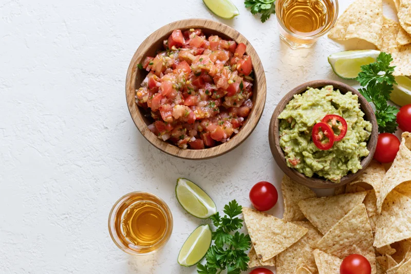 Mexican appetizer spread with salsa, guacamole, tortilla chips and tequila on a white textured surface
