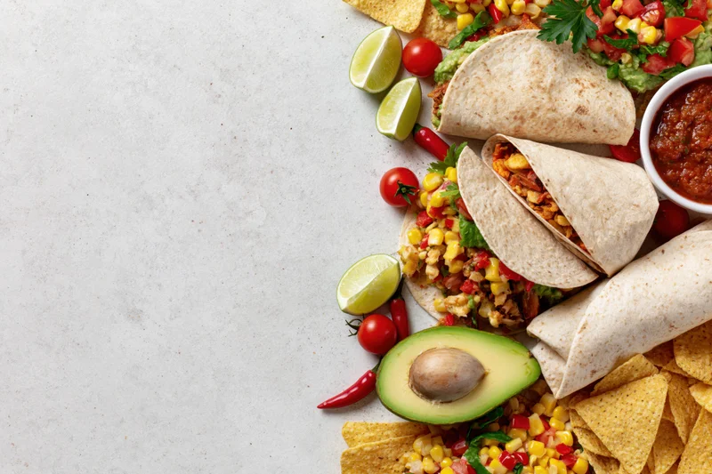 Mexican food spread with tacos, avocado, salsa, and chips arranged along the right edge of a white surface