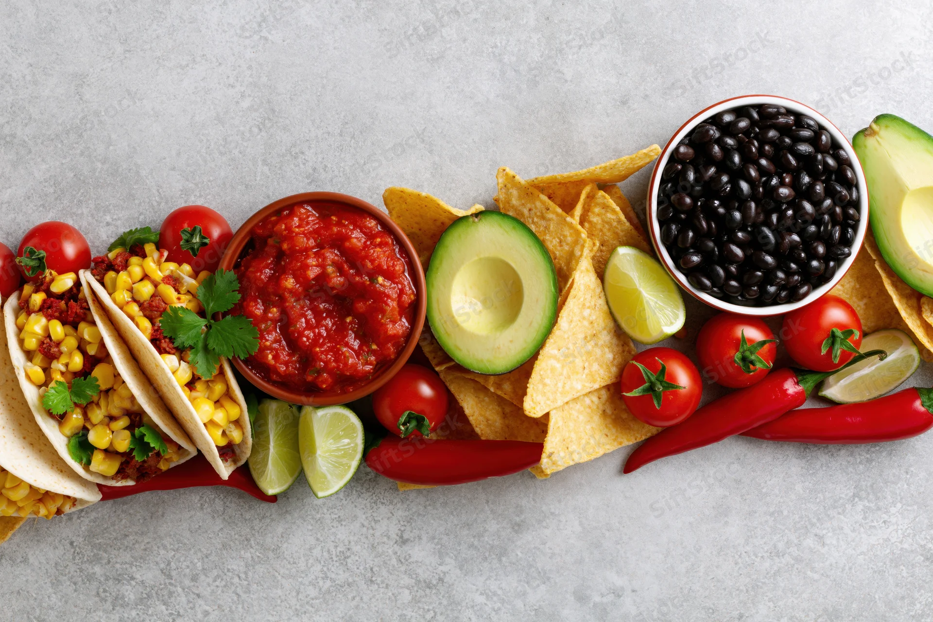 Mexican food spread with tacos, salsa, avocado, black beans, and chips arranged along bottom edge of frame on gray surface