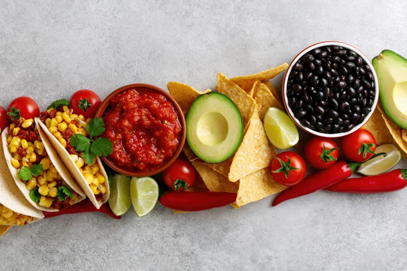 Mexican food spread with tacos, salsa, avocado, black beans, and chips arranged along bottom edge of frame on gray surface