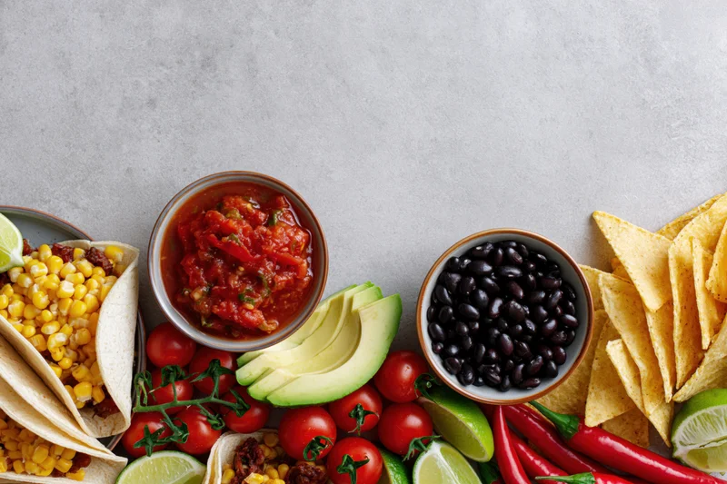 Mexican food spread laid flat along bottom edge of frame with tacos, salsa, avocado, black beans, chips, and vegetables on gray surface
