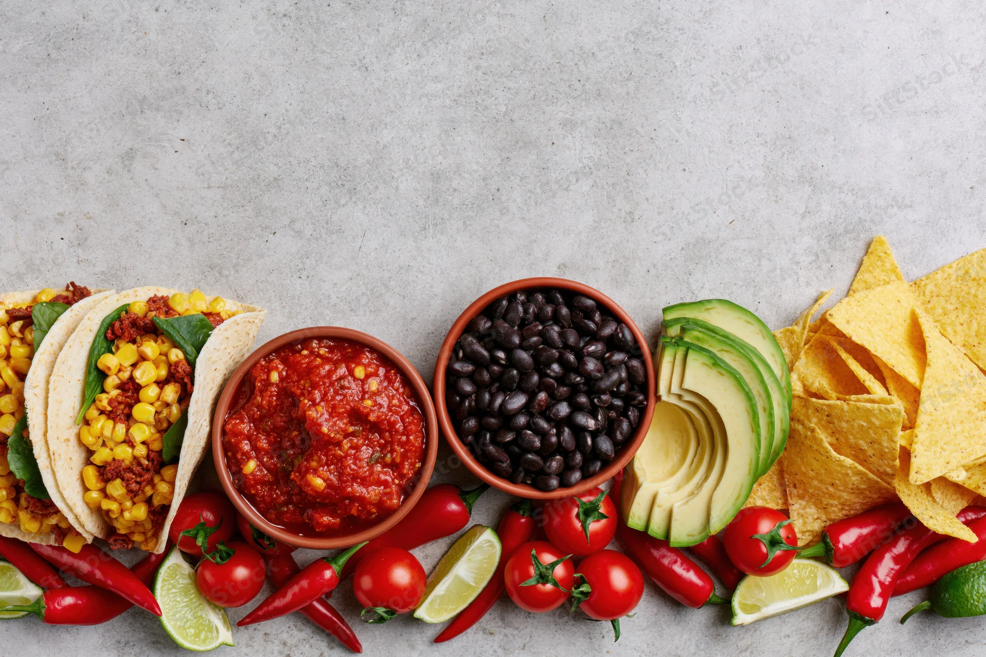 Mexican food spread laid flat along bottom edge of frame with tacos, salsa, black beans, avocado, chips, chili, lime, and tomato on gray surface