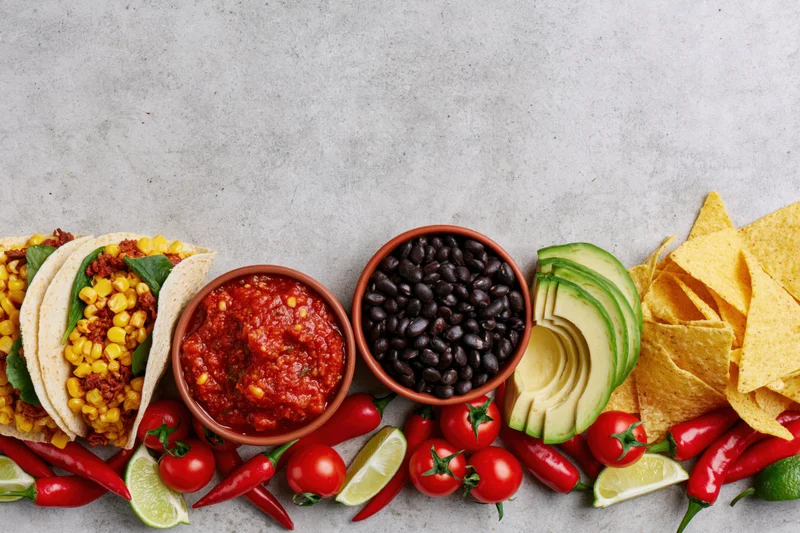 Mexican food spread laid flat along bottom edge of frame with tacos, salsa, black beans, avocado, chips, chili, lime, and tomato on gray surface