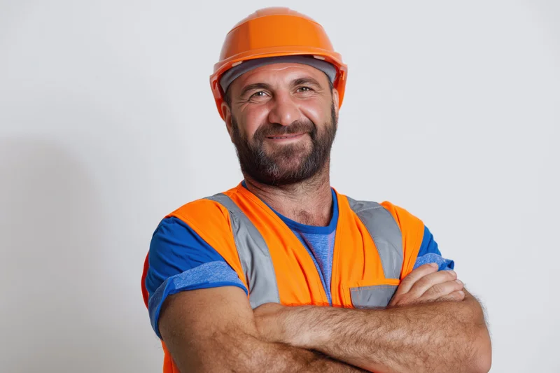 Middle-aged construction worker with arms crossed smiling in orange safety helmet and vest against white background