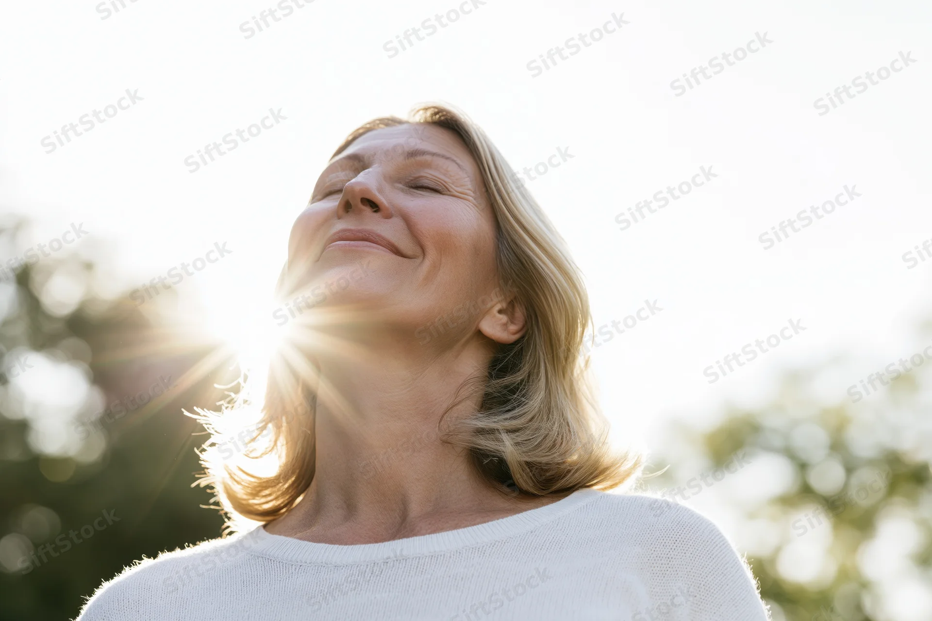 Middle-aged woman smiling with eyes closed, tilting head back into sunlight outdoors