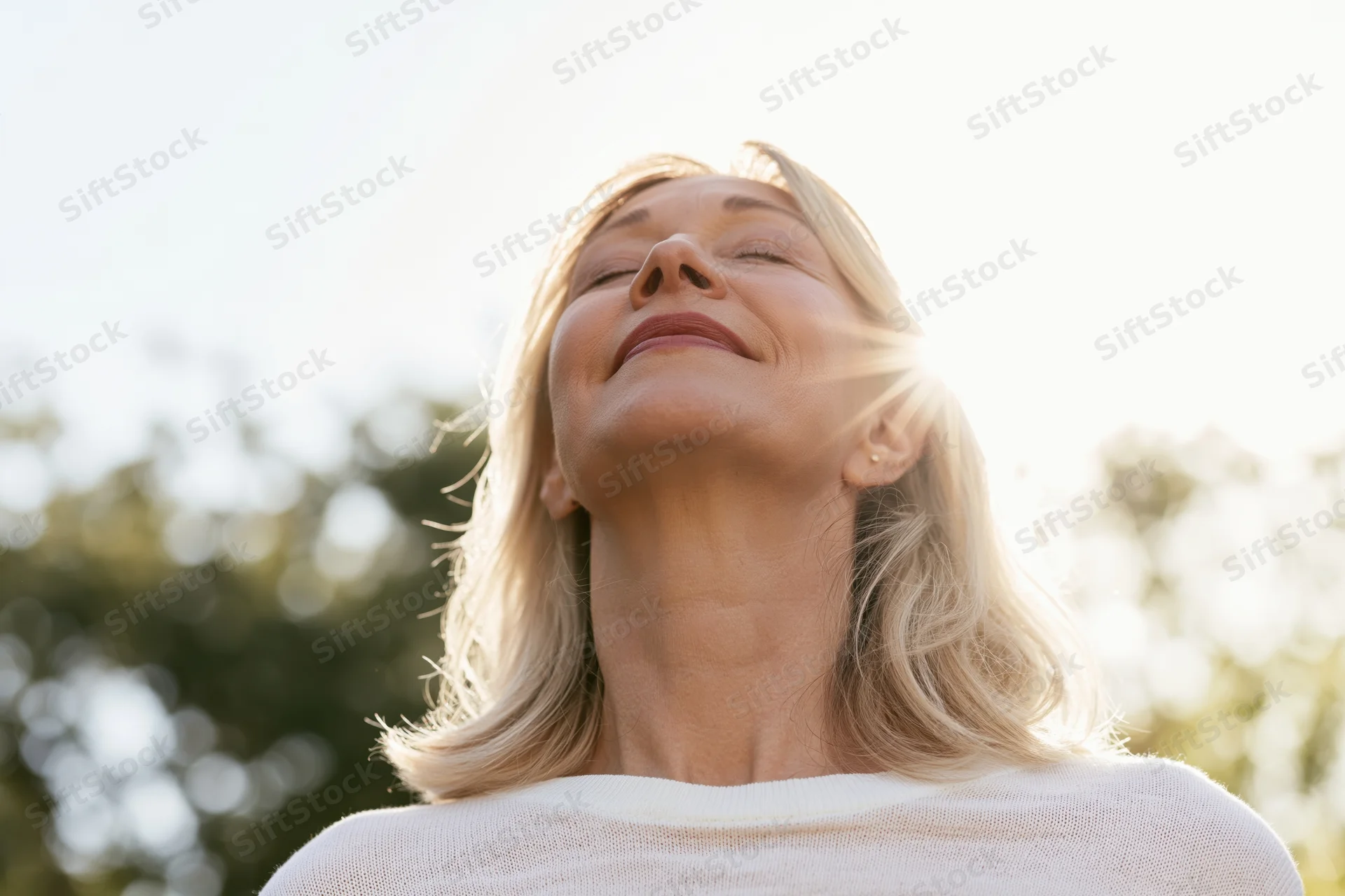Middle-aged woman with shoulder-length blonde hair tilting head back, eyes closed, smiling in sunlight outdoors