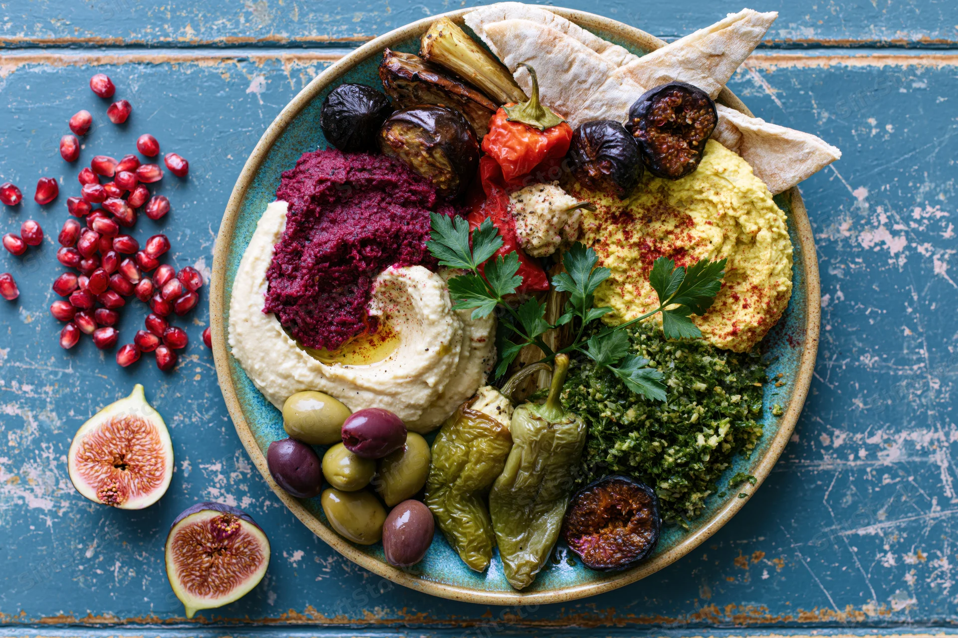 Overhead view of a middle eastern mezze platter with hummus, beetroot dip, pita, olives, figs and pomegranate on a blue wooden table
