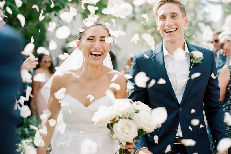 Bride and groom laughing as they walk through falling white flower petals at outdoor wedding ceremony