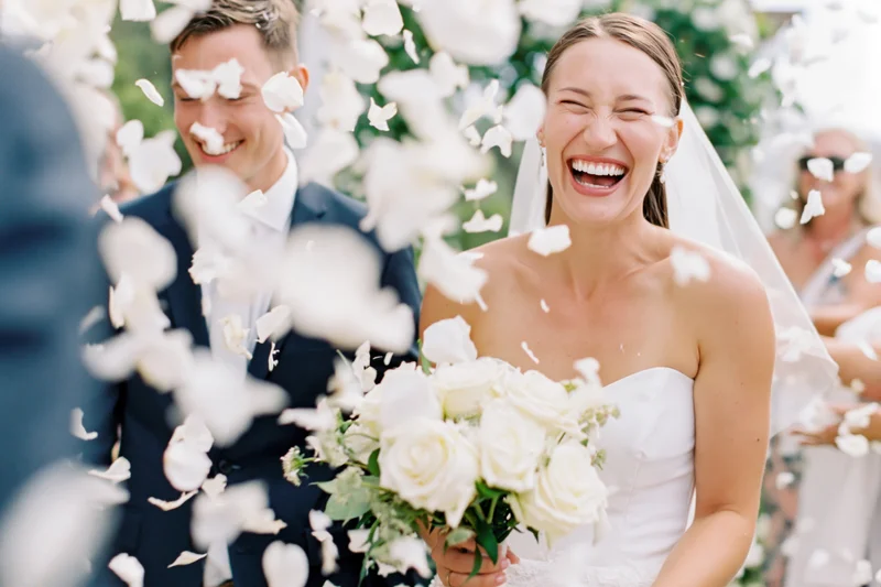 Bride and groom laughing as flower petals fall around them outdoors