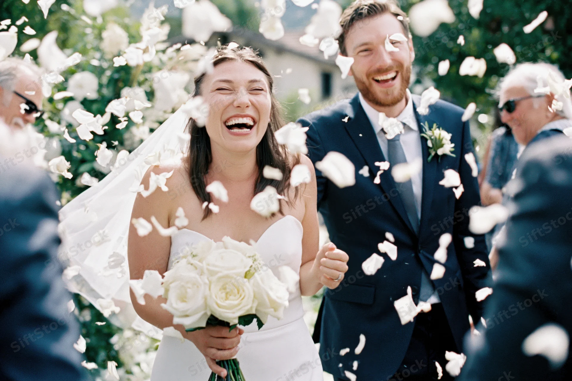Bride and groom laughing as guests toss white flower petals around them outdoors