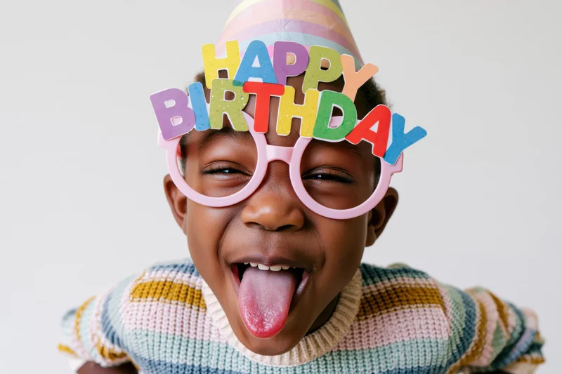 Child wearing colorful happy birthday glasses and party hat, sticking tongue out, in studio setting