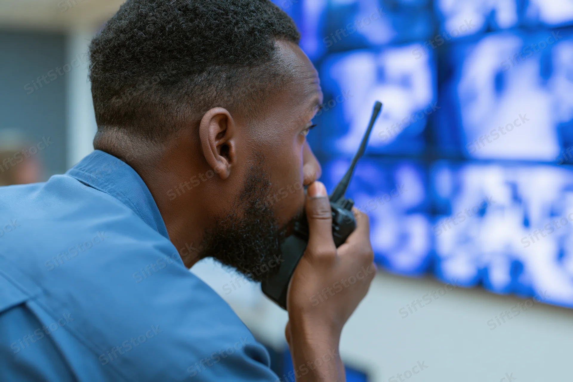 Man in blue uniform speaking into walkie-talkie in front of surveillance monitors
