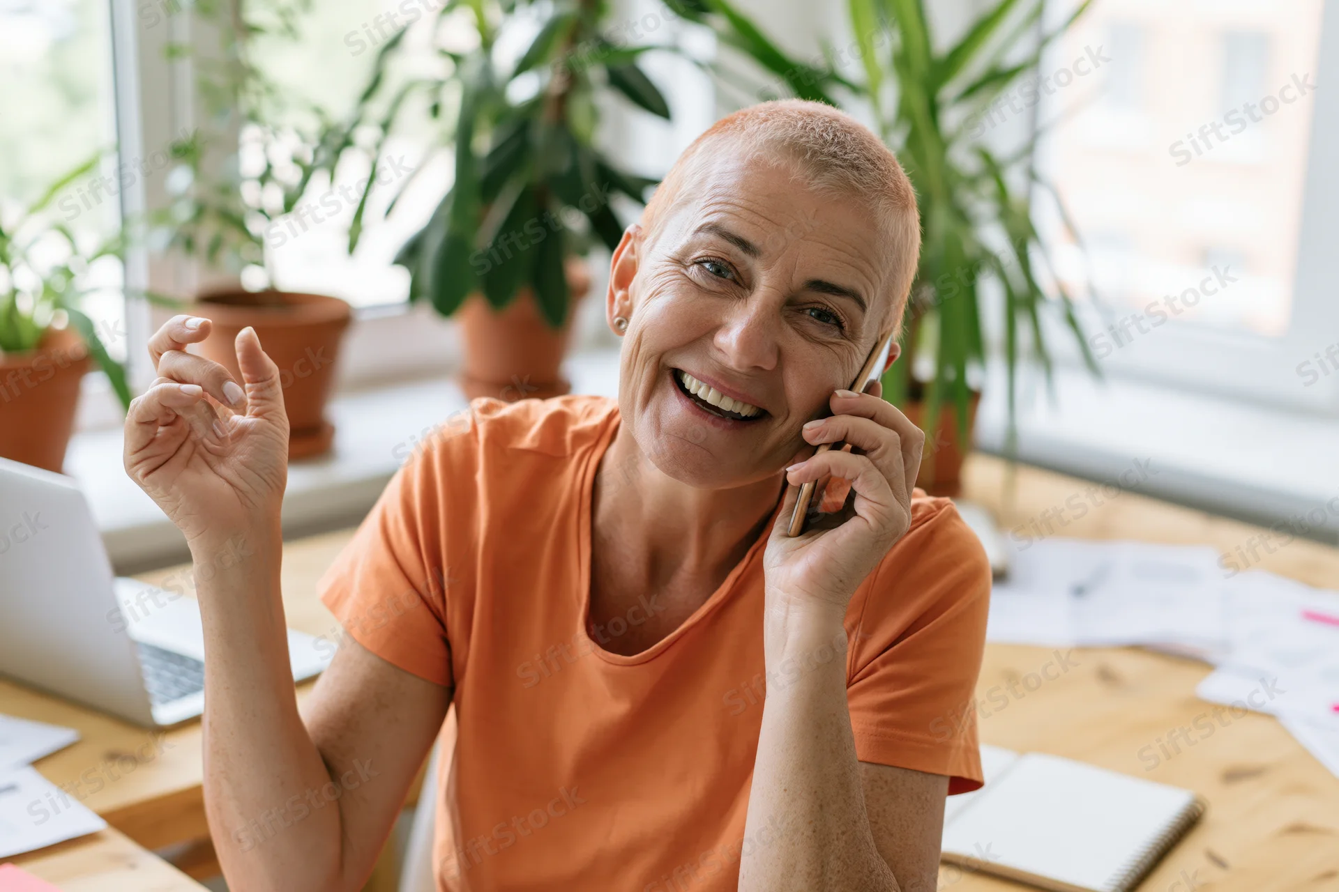 Smiling woman talking on phone at desk with laptop and plants in home office