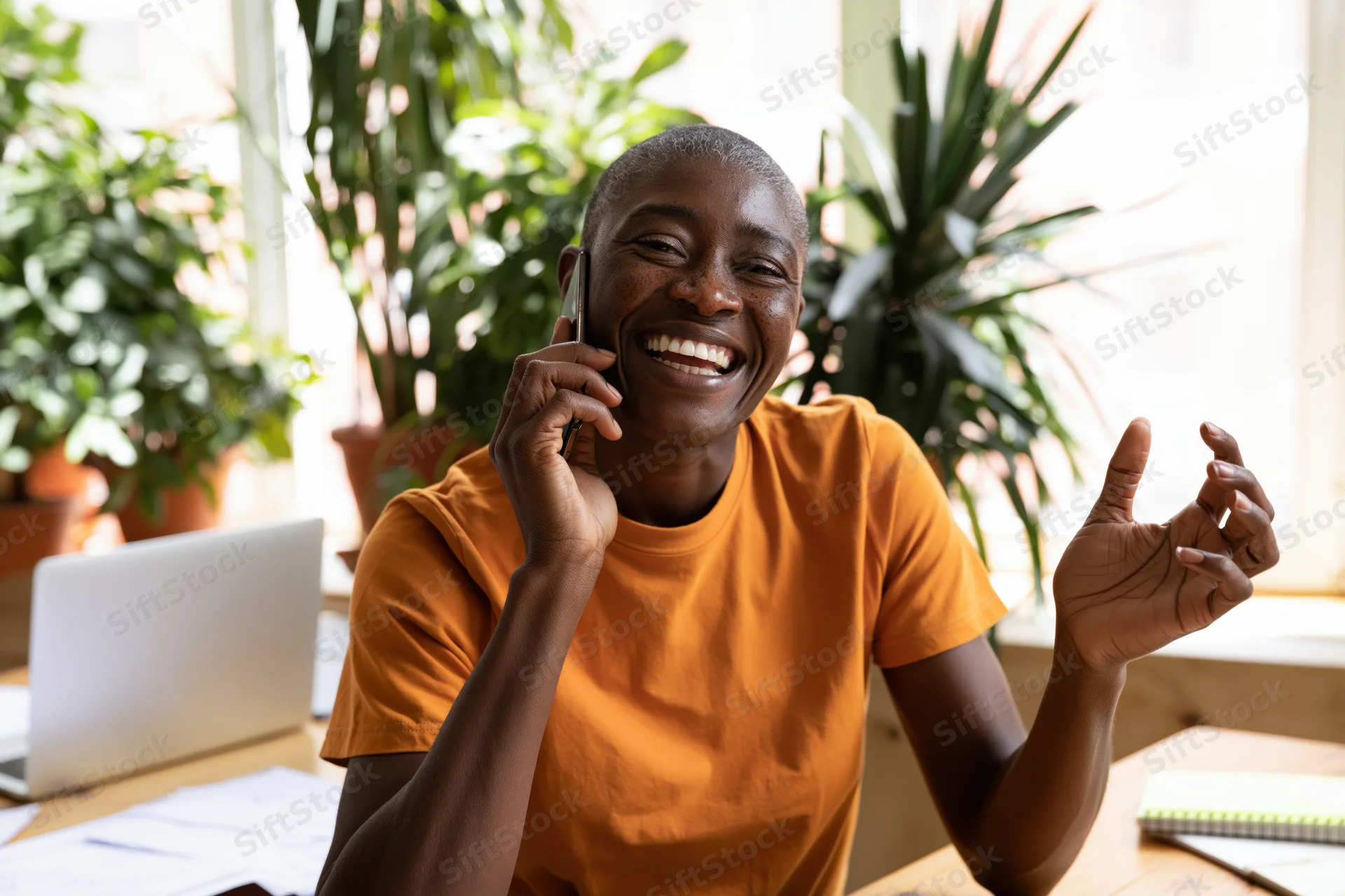 Woman smiling while talking on phone at desk with laptop and plants in office