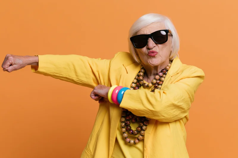 Senior woman dabbing in yellow jacket and sunglasses against orange background