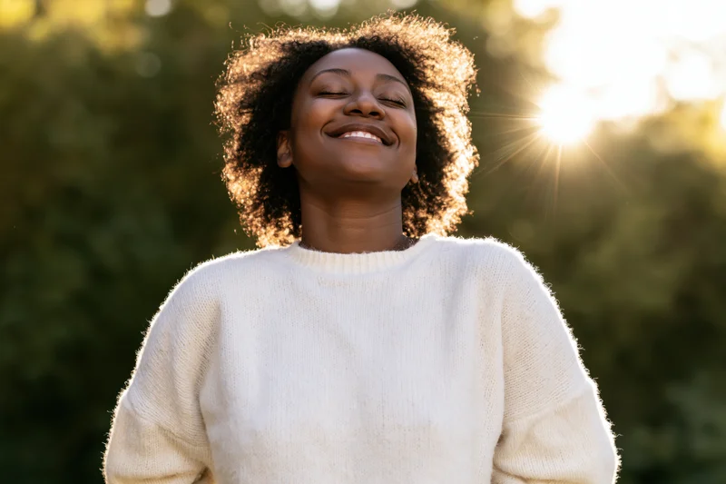 Woman smiling with eyes closed outdoors in sunlight wearing cream sweater