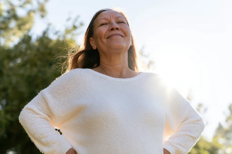 Senior woman smiling with eyes closed in sunlight outdoors, hands on hips, surrounded by trees