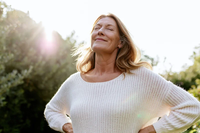 Senior woman standing outdoors with eyes closed and hands on hips, basking in sunlight among greenery
