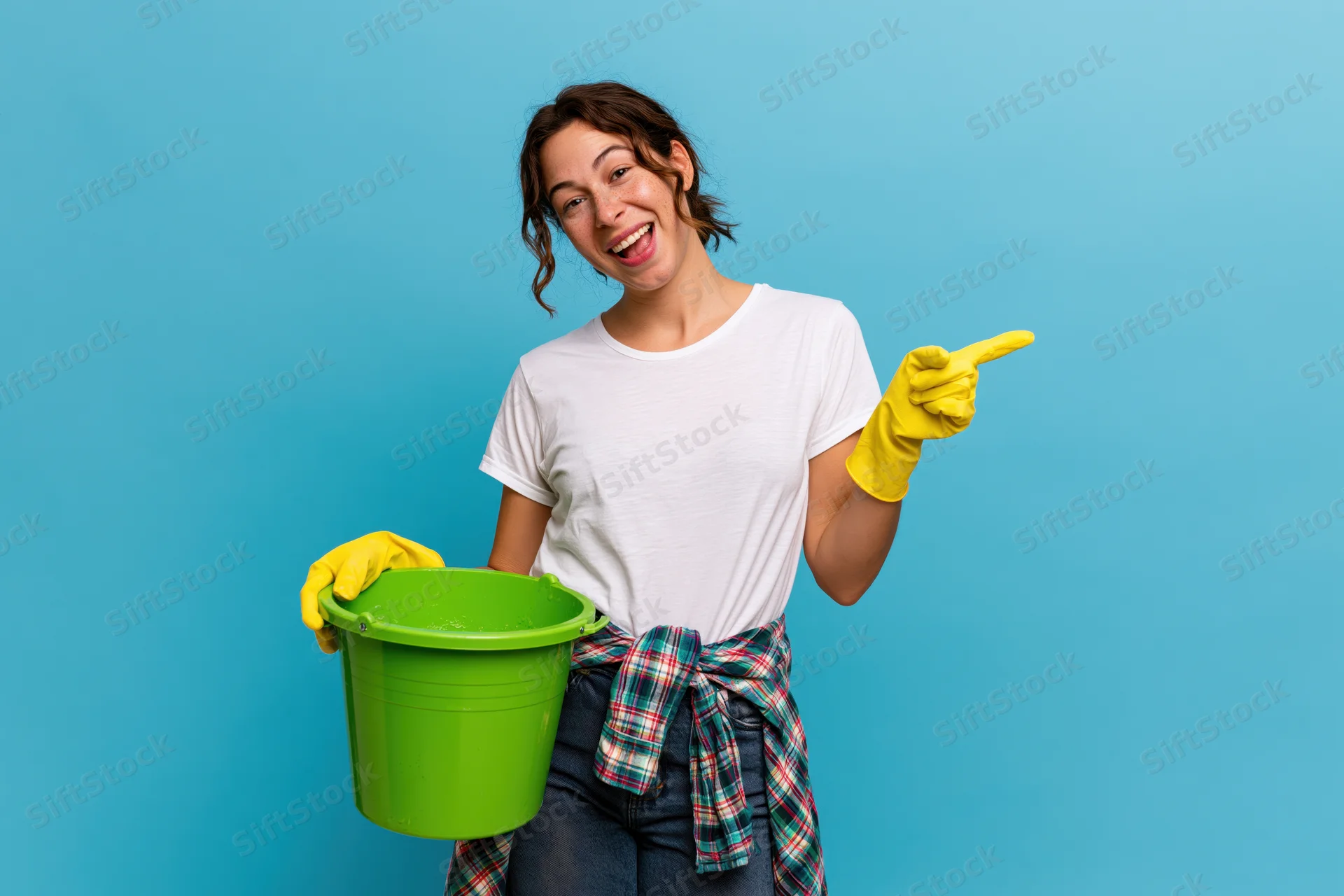 Woman holding green bucket and wearing yellow gloves points to side while smiling against blue background