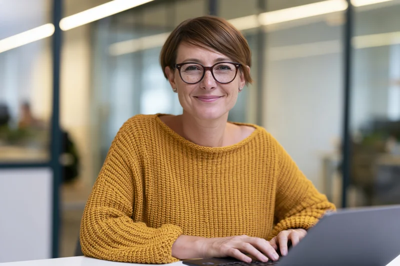 Woman smiling at camera while typing on laptop at office desk