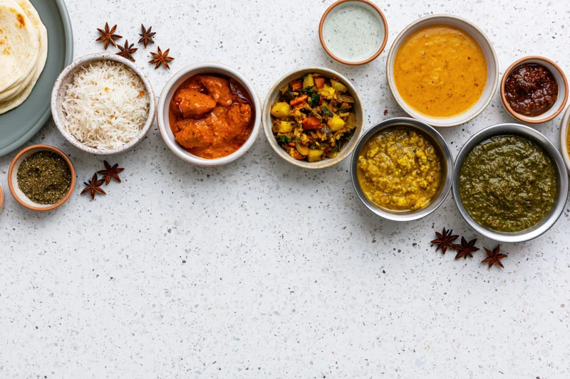 Overhead flat-lay of assorted indian dishes arranged on a white countertop with bowls of curry, rice, naan, dal, chutney, and spices
