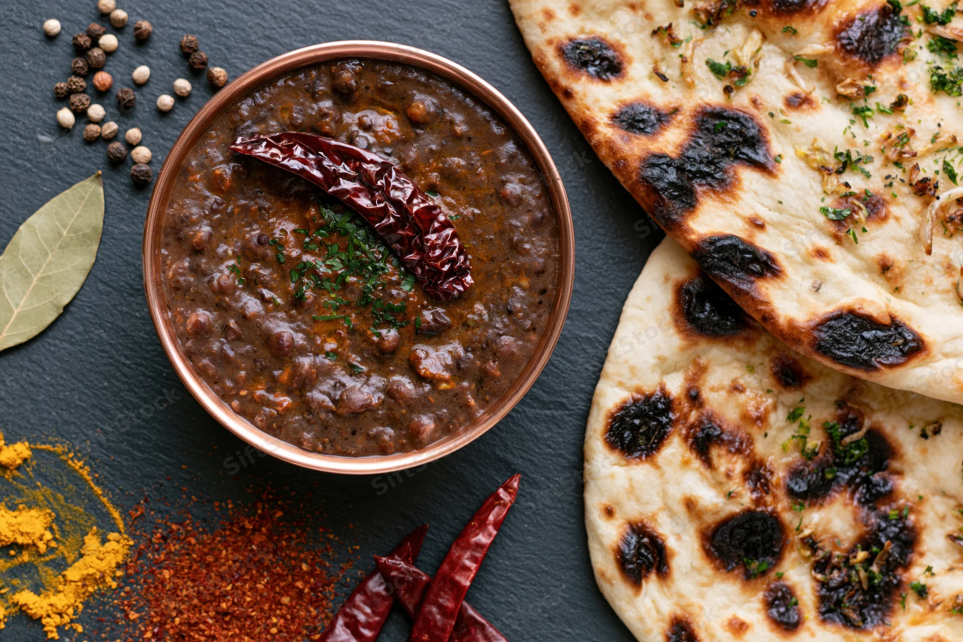 Copper bowl of dal makhani with naan bread and spices arranged on dark slate surface