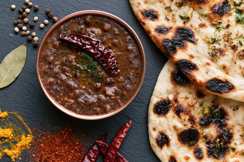 Copper bowl of dal makhani with naan bread and spices arranged on dark slate surface